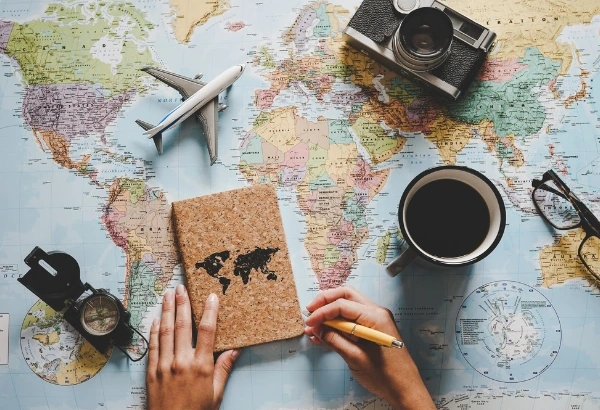World map and travel items on a tabletop: model airplane, camera, notebook, hands holding a pencil