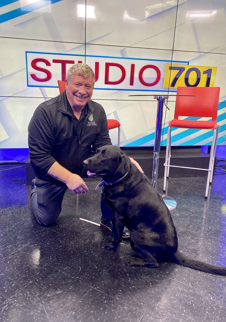 Man kneeling beside a black Labrador retriever on a television studio set with a 'STUDIO 701' sign