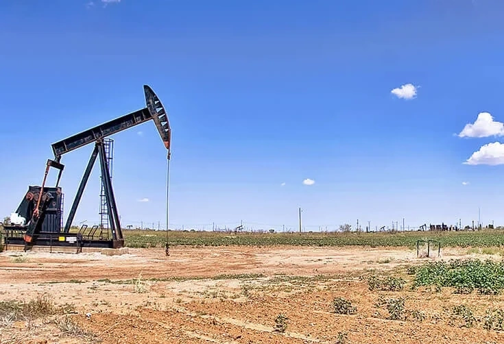 Black oil pumpjack standing in a dusty field under a clear blue sky, representing oil and gas operations