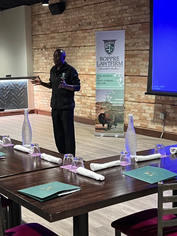 Speaker in a brick conference room beside a law firm banner; tables set with folders and water for a legal workshop