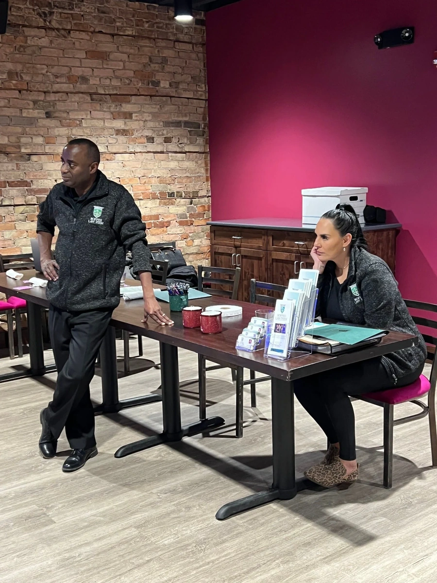 Man and woman at a table with brochures, boxes, and office materials against a brick and magenta wall