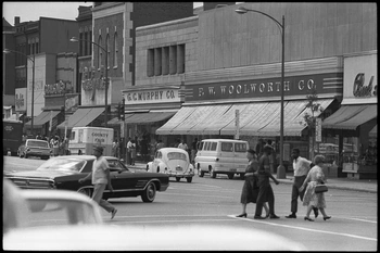People crossing a busy main street lined with neighborhood shops.