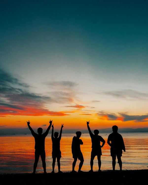 Silhouettes of five people with their arms raised, standing on a beach at sunset. The sky is filled with vibrant shades of orange, red, and purple, reflecting off the calm waters.