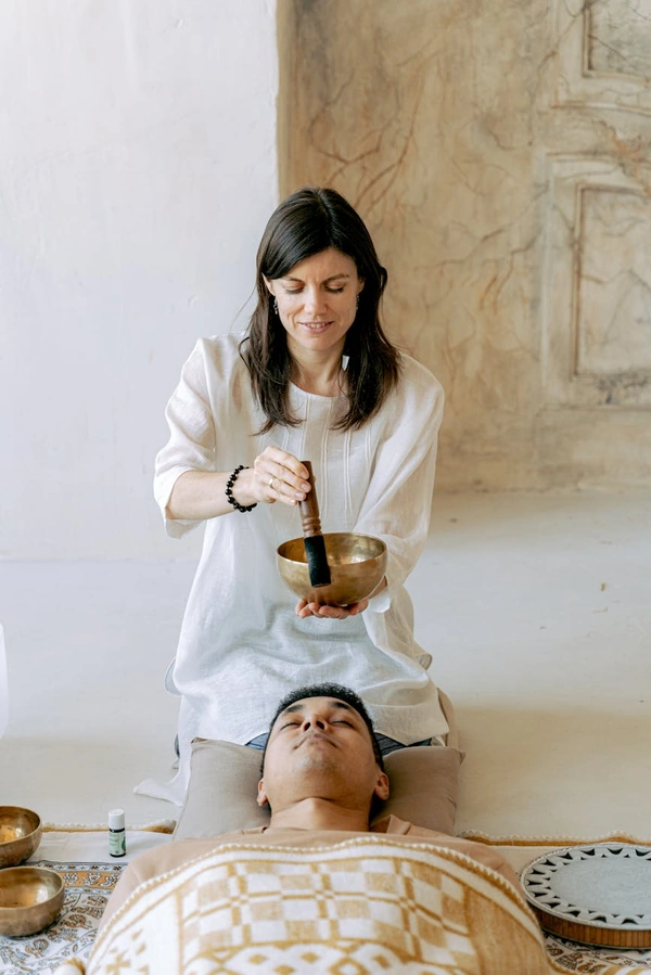 A woman plays a Tibetan singing bowl during a healing session with a man lying peacefully.