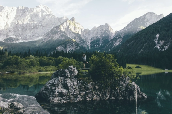 Woman doing a yoga pose on a rock island in the middle of a lake with large surrounding mountains