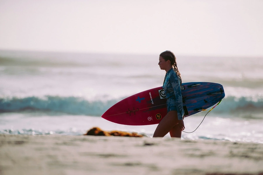 Surfer walking her surfboard down the beach