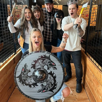 Enthusiastic group at a Viking-themed axe throwing venue, posing with props including a decorative Viking shield with Norse design, battle axes, and costume crowns in an indoor wooden lane