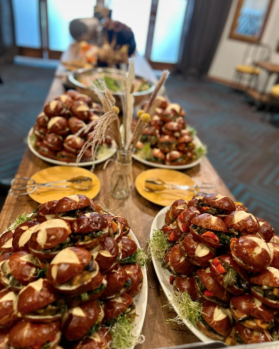 Elegant catering display of gourmet pretzel sandwiches on white platters, arranged on a wooden table with yellow plates. Each pretzel sandwich is filled with fresh vegetables and garnished with microgreens, featuring distinctive cross-hatched patterns on the glossy brown pretzel buns.