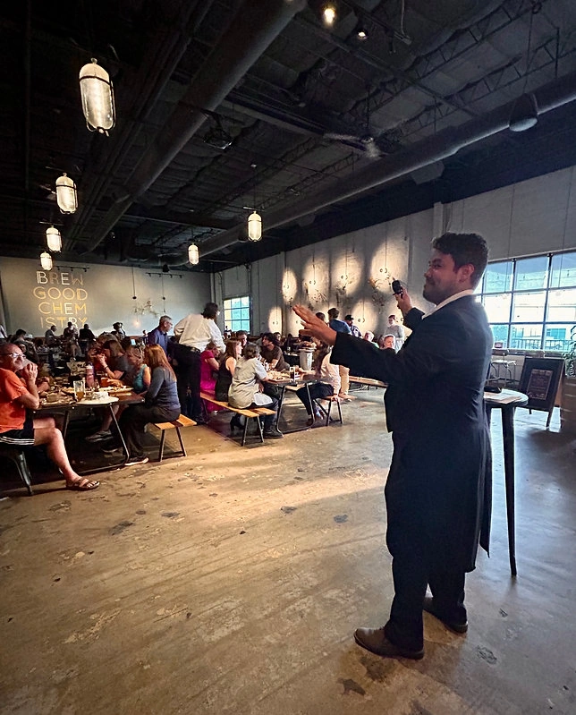 A person in a dark suit speaking to a packed restaurant crowd, with industrial pendant lights hanging from an exposed ceiling and "BE GOOD CHEM" signage visible on the wall