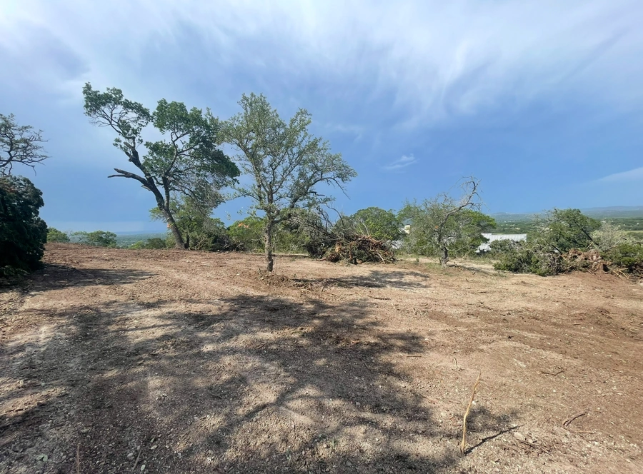 A cleared hillside with scattered oak trees against a blue sky, showing signs of recent land development or clearing with exposed dirt ground and some remaining vegetation