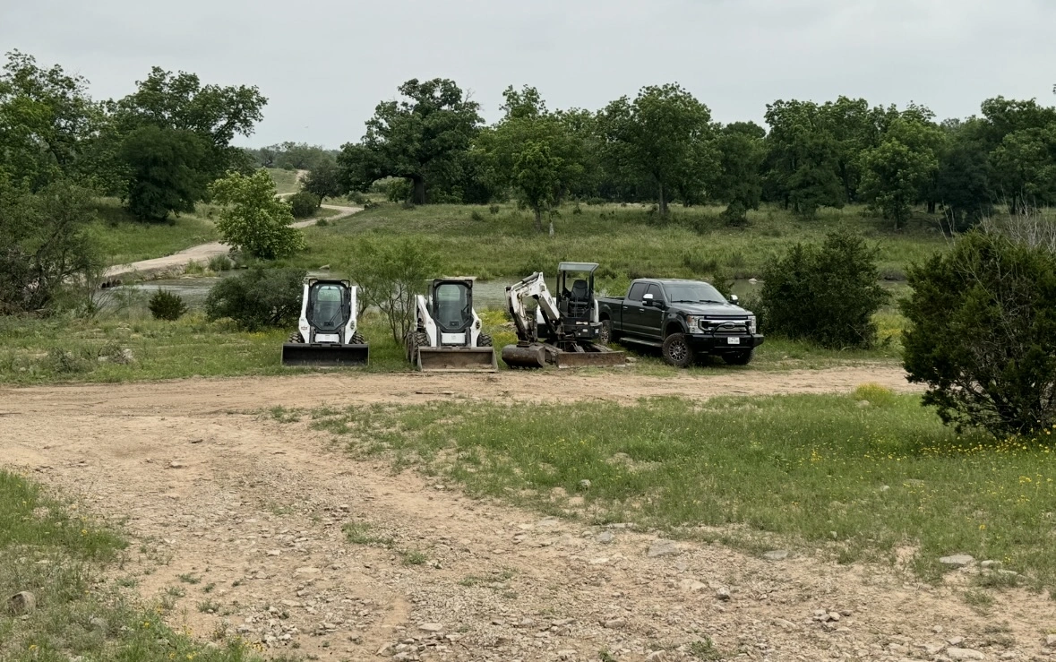 Three compact construction machines (two Bobcat skid steers and one mini excavator) lined up next to a black Ford pickup truck against a backdrop of rolling hills and oak trees on a rural dirt path