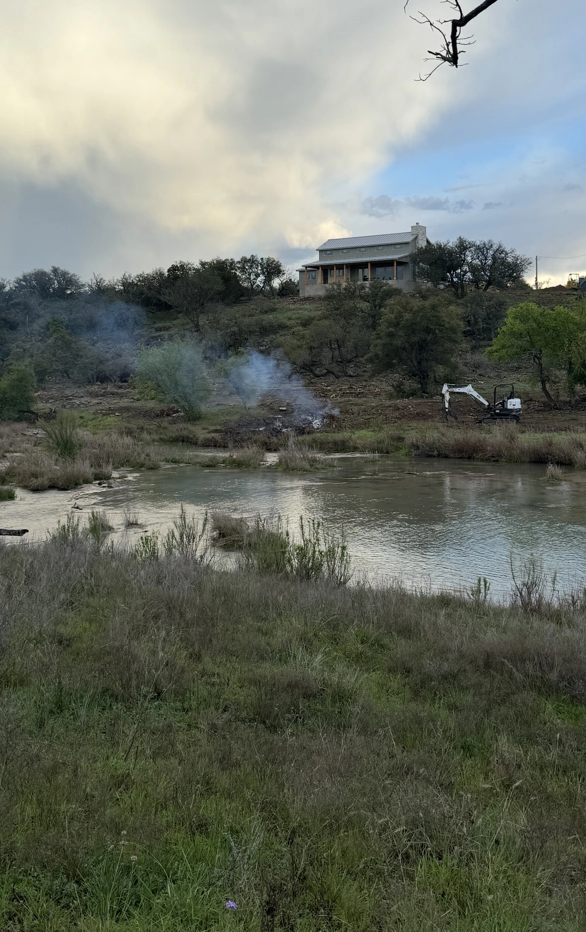 Modern farmhouse-style home perched on a rugged Texas Hill Country hillside overlooking a serene pond, with wisps of smoke from a controlled burn and a small excavator visible in the landscape