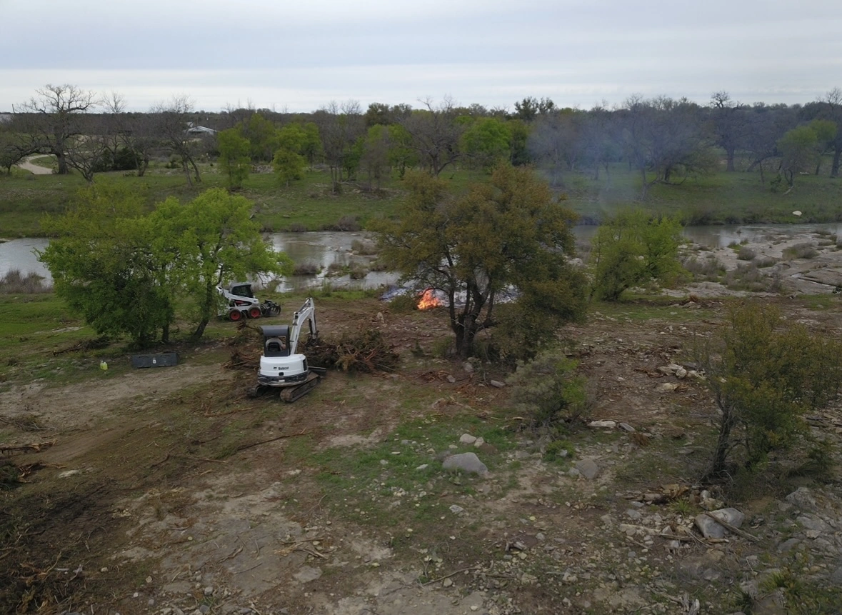 Aerial photograph showing land development work with a white mini excavator clearing terrain near a riverbank, featuring scattered trees, rocks, and a small controlled burn with rising smoke