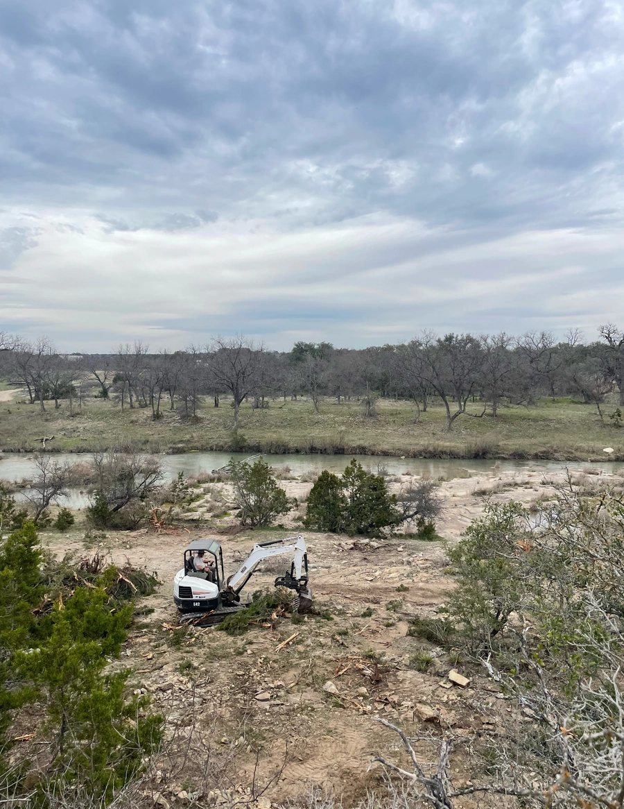 Mini excavator working on a rocky riverbank with bare winter trees, overlooking a winding river in Texas Hill Country under cloudy skies