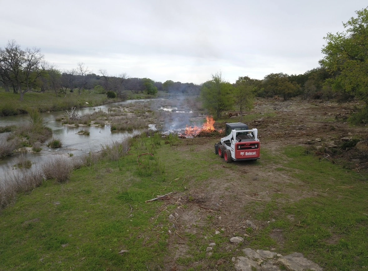 Controlled brush burning operation along a riverbank with a Bobcat skid-steer loader parked nearby, showing land clearing and habitat management activities in a natural setting