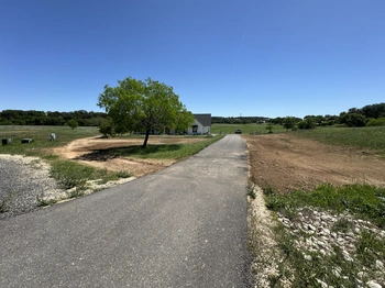 Long paved driveway leading to a white farmhouse with grey roof, surrounded by Texas Hill Country landscape with green mesquite trees and natural grassland under bright blue sky