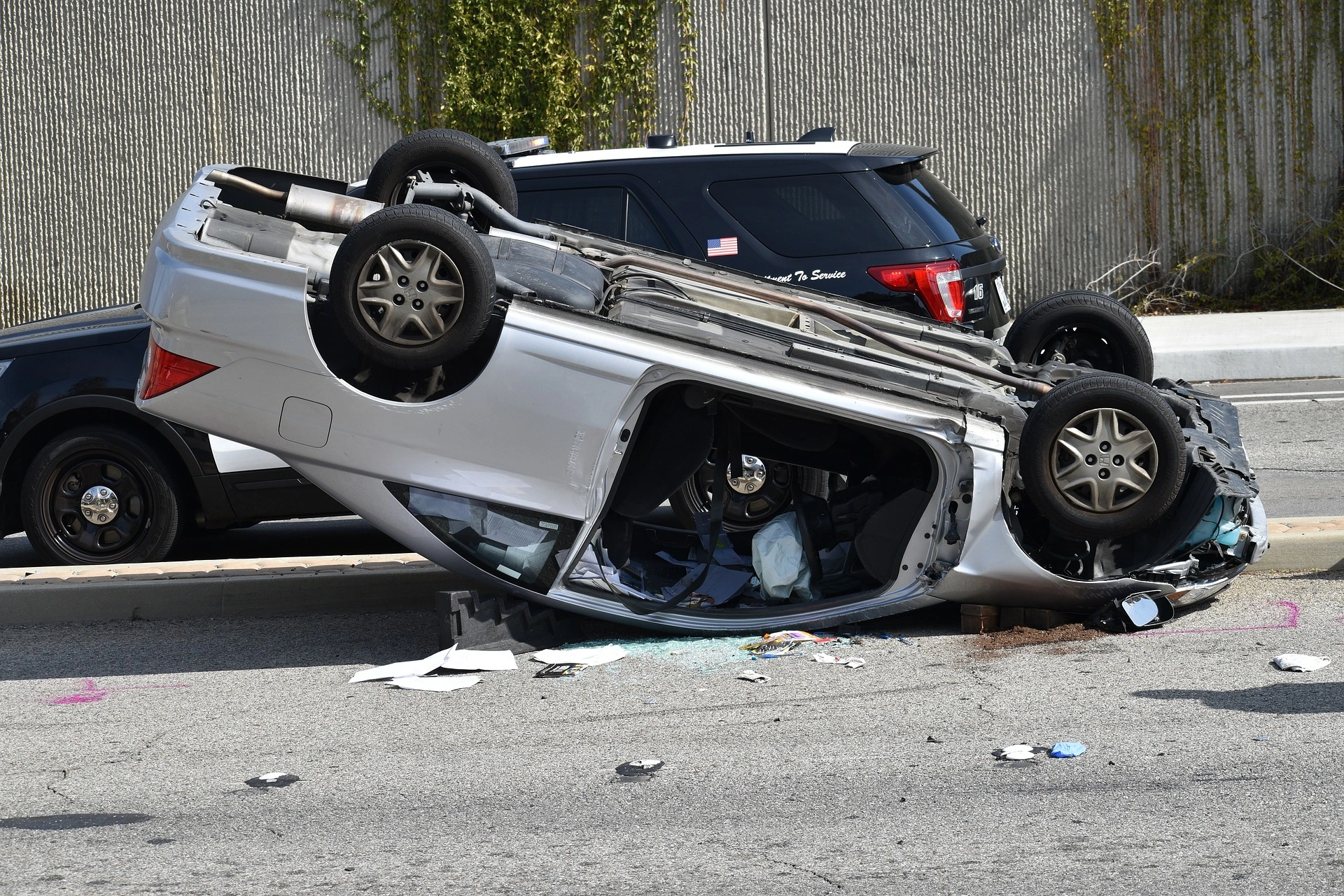 An overturned car on its side, with debris scattered on the ground around it. The car appears to be a silver or grey sedan. The scene suggests a serious automobile accident has occurred.