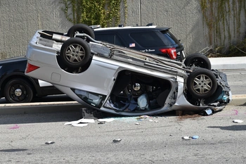 An overturned car on its side, with debris scattered on the ground around it. The car appears to be a silver or grey sedan. The scene suggests a serious automobile accident has occurred.