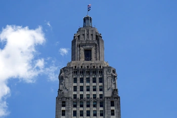 The image shows a tall, ornate stone skyscraper with intricate architectural details and a prominent spire at the top. The building stands against a blue sky with fluffy white clouds, conveying a sense of grandeur and stateliness.