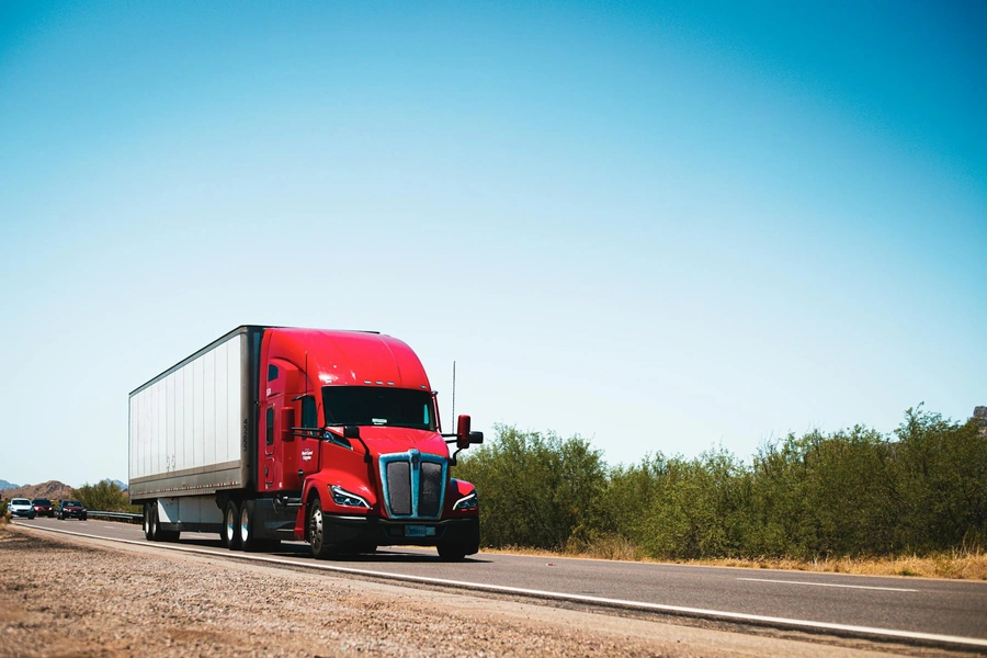 A large red semi-truck hauling a cargo trailer drives down a rural highway with green trees and a blue sky in the background.